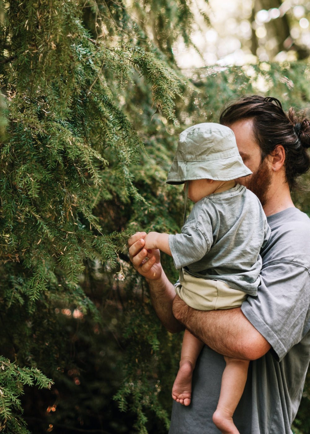 Man holding a baby outside touching a pine tree
