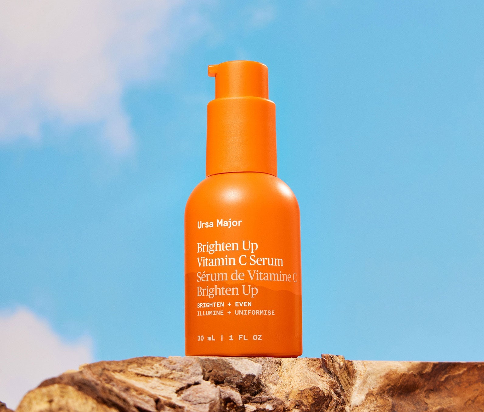 Orange bottle of Ursa Major Brighten Up Vitamin C Serum on a wooden surface with a blue sky background