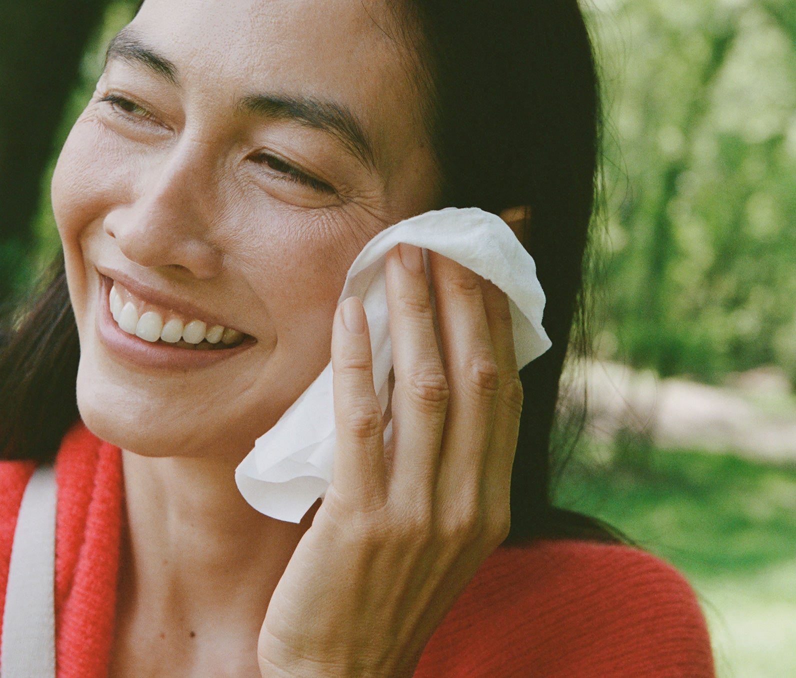 Smiling woman cleaning her face with a white facial wipe in a wooded, outdoor setting