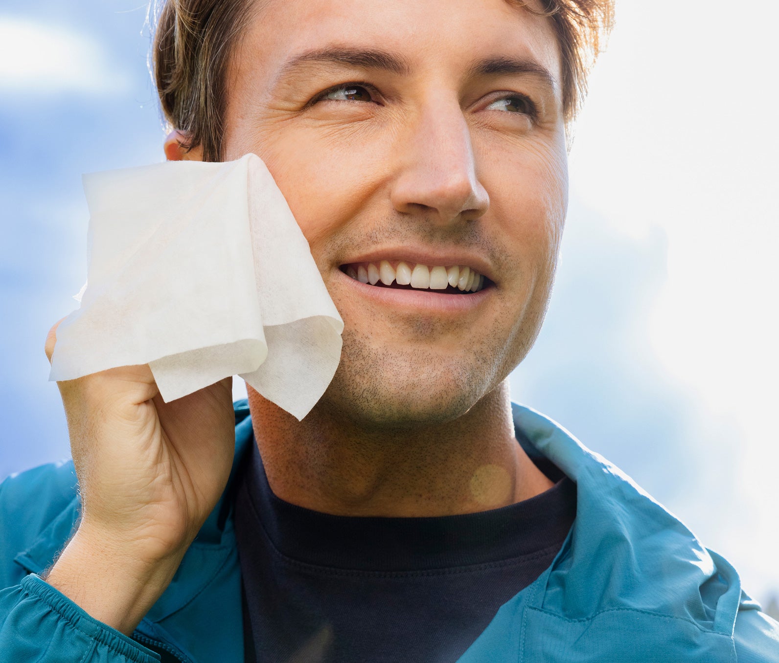 Man cleaning his face with a white facial wipe against a blue sky background