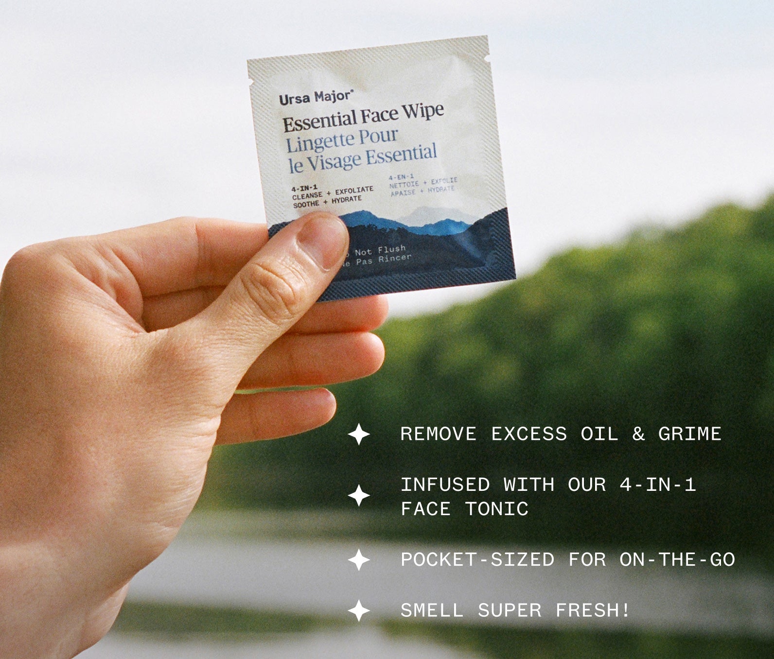 Hand holding a Ursa Major Essential Face Wipe against a blurred, lakeside background, with text about product features, including: Remove excess oil and grime; Infused with our 4-in-1 Face Tonic; Pocket-sized for on-the-go; Smell super fresh!