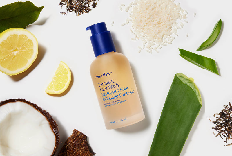 Glass Ursa Major face wash bottle surrounded by lemons, coconuts, and leaves on a white background