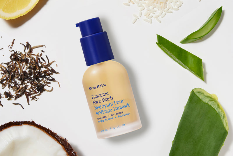 Ursa Major Fantastic Face Wash bottle with a blue pump, styled in a flat lay with natural ingredients like aloe vera, rice, tea, and coconut on a white background.