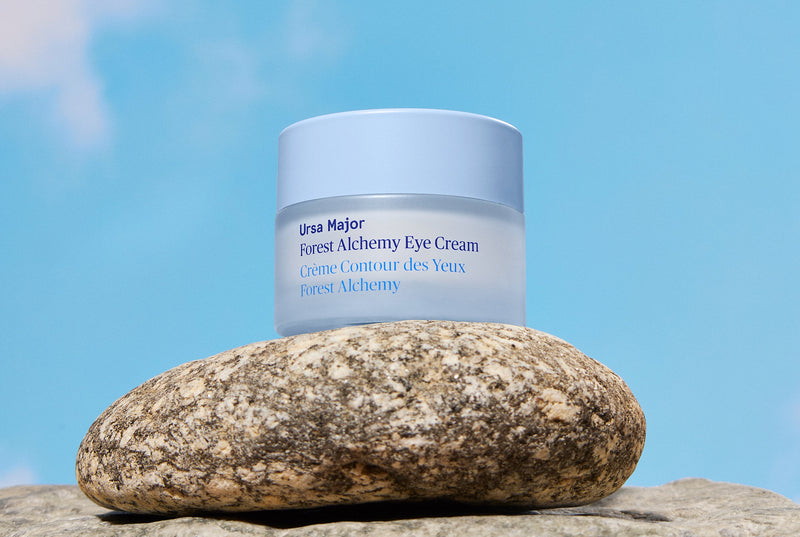 Blue jar of Forest Alchemy Eye Cream resting on stone surface amidst a clear, blue sky