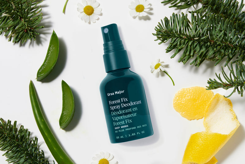 Green bottle of forest fix spray deodorant surrounded by green leaves, flowers, and lemon peel on a white background