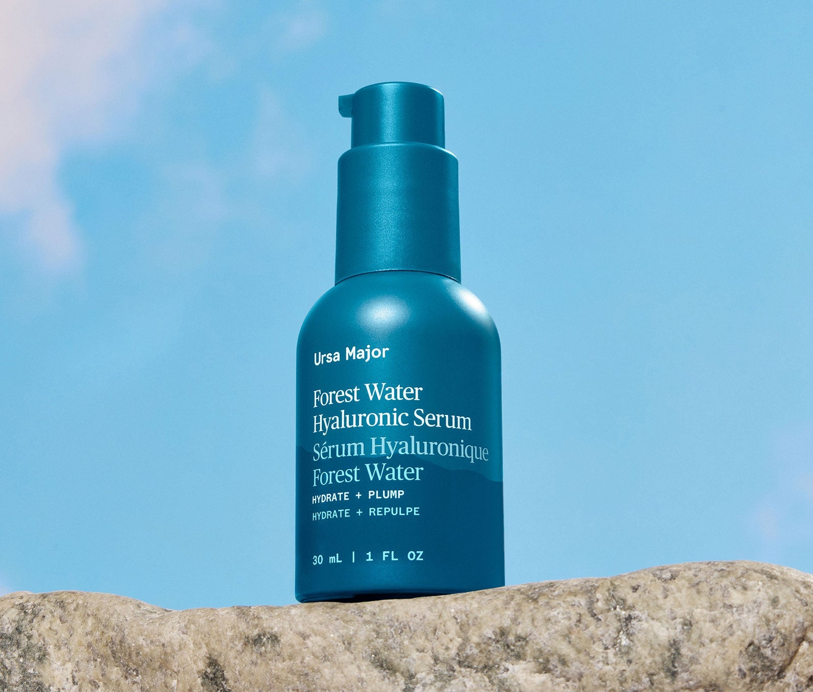 Blue bottle of Ursa Major Forest Water Hyaluronic Serum on a rock with a blue sky background