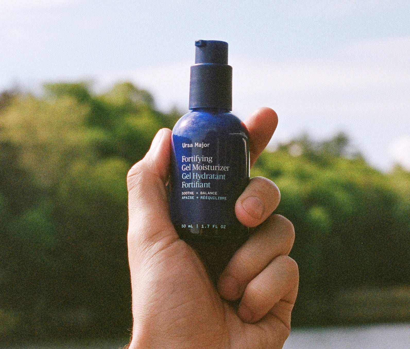 Hand holding a blue bottle of Ursa Major Fortifying Gel Moisturizer with a blurred forest background