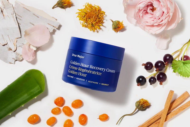 Blue jar of Ursa Major Golden Hour Recovery Cream surrounded by flowers and herbs on a white background