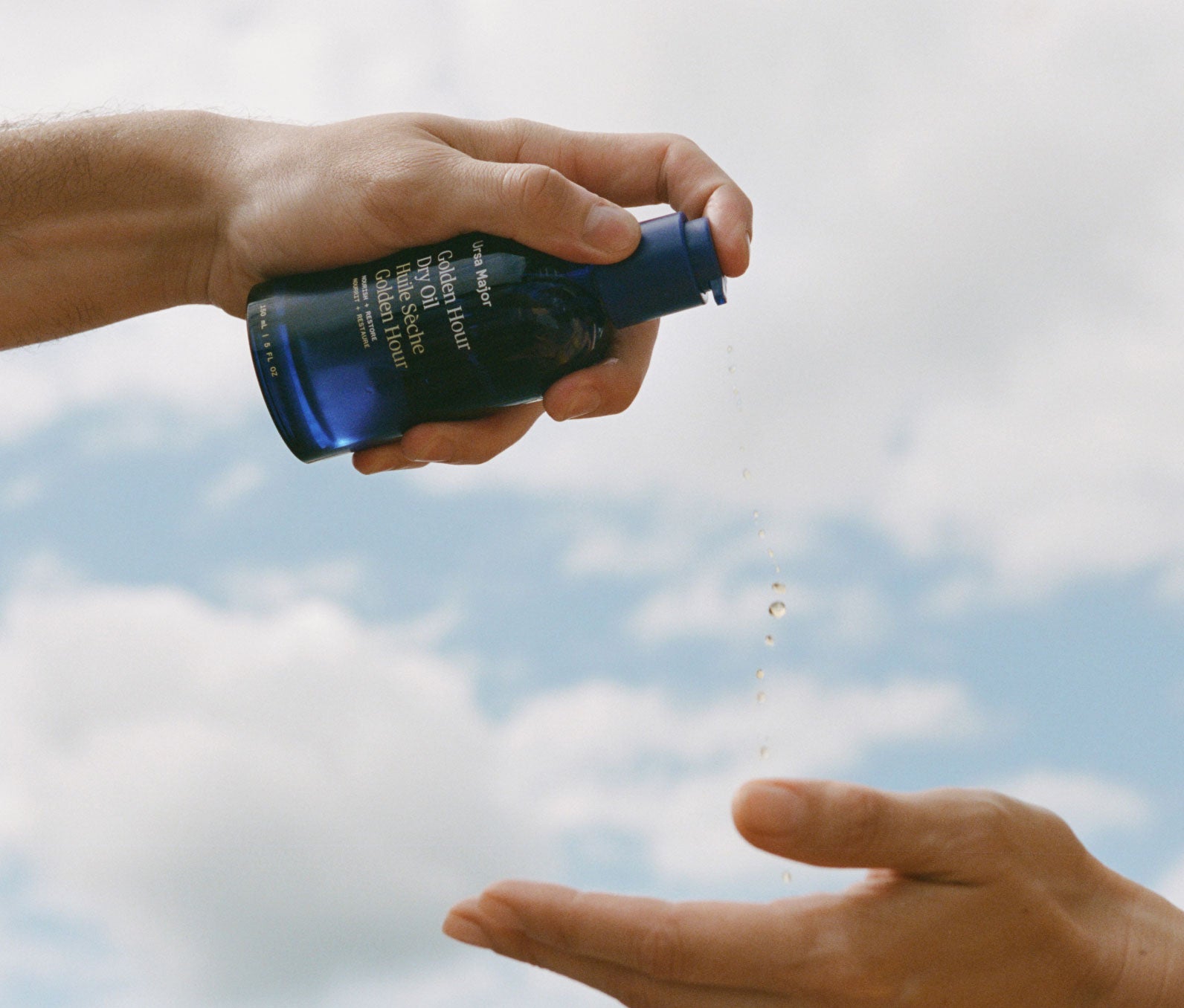 Person dispensing drops of Golden Hour Dry Oil from blue bottle into an open palm against a cloudy sky