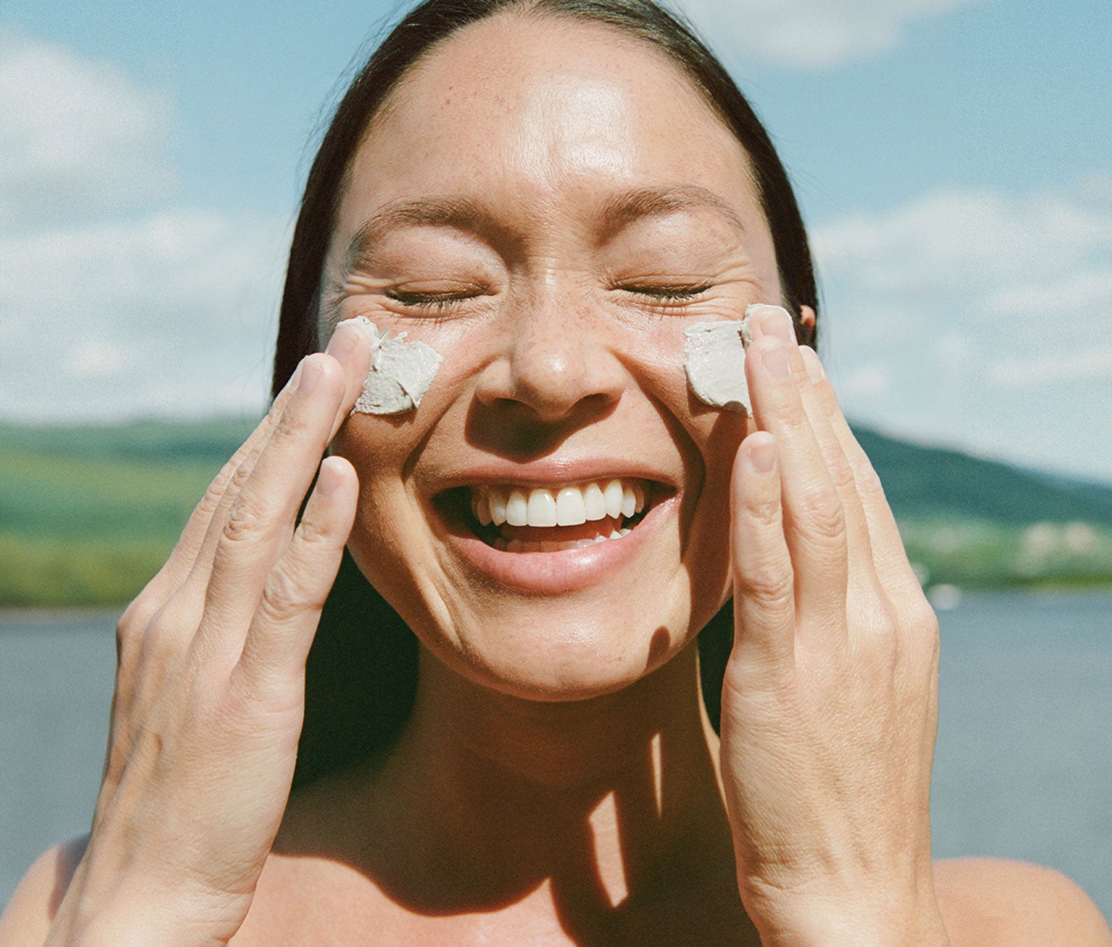 Smiling woman applying mineral polish to her cheeks with a scenic, lakeside background