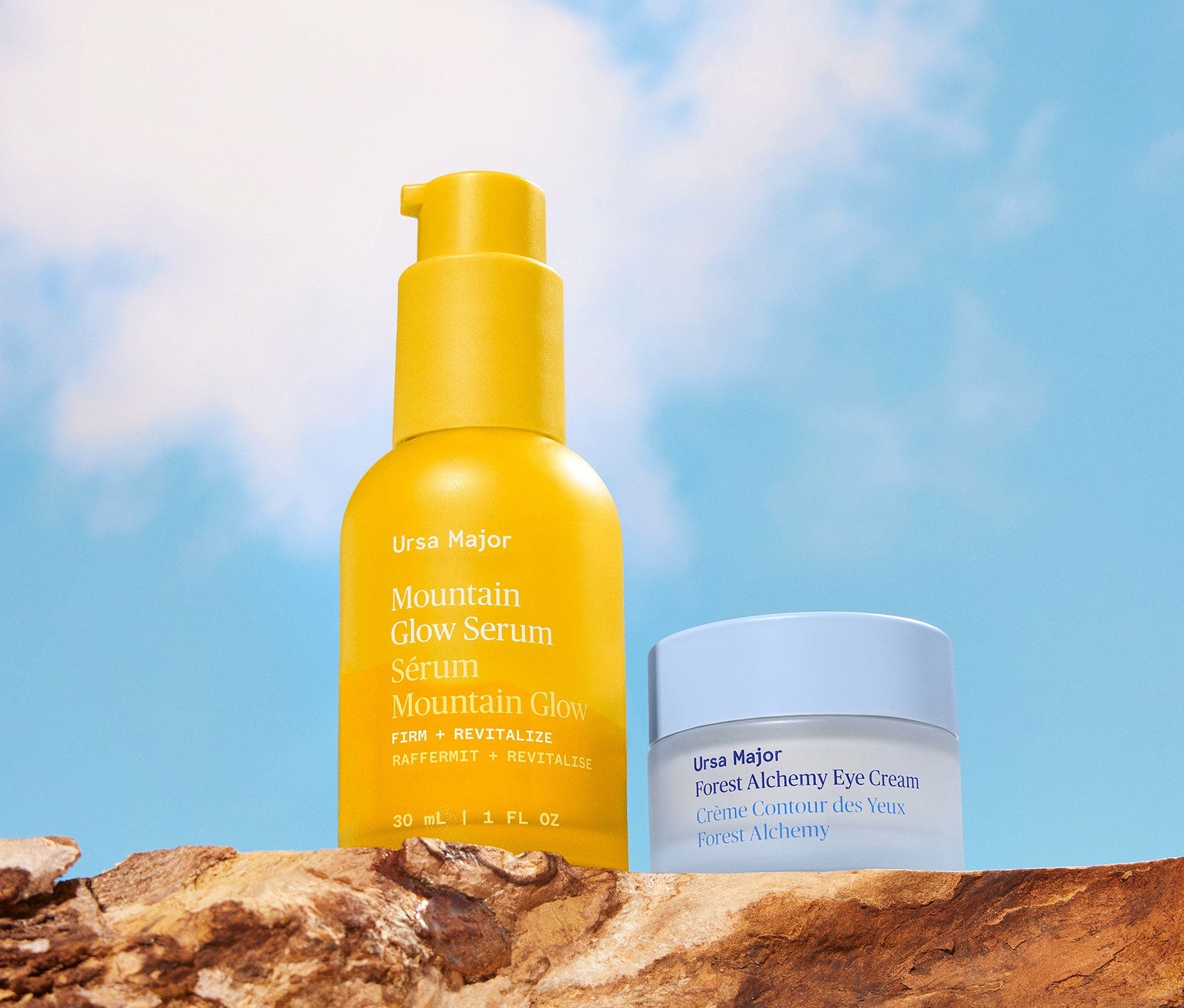 Yellow bottle of Mountain Glow Serum next to a blue jar of Forest Alchemy Eye, on a wooded surface with a blue sky background