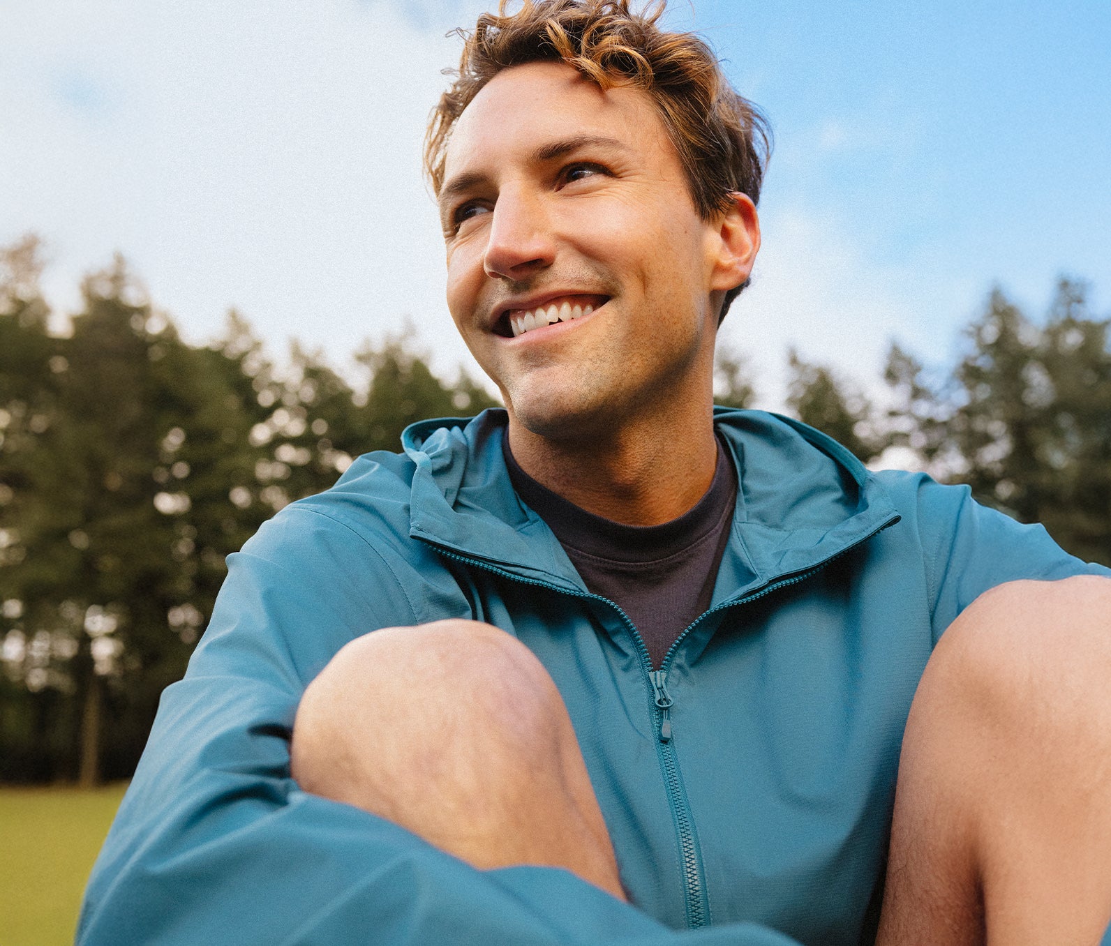 Person sitting outside in green grass smiling into the distance wearing a blue rain jacket.