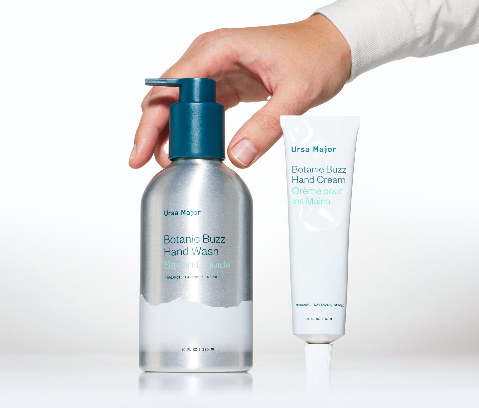 Aluminum bottle of Ursa Major Botanic Buzz Hand Wash and tube of Ursa Major Botanic Buzz Hand Cream on a white surface with a white background. Hand is placing the hand wash onto the surface.