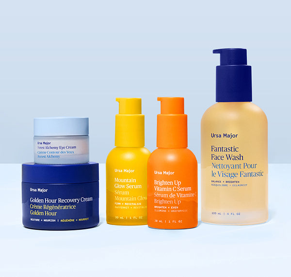 Collection of Ursa Major skincare products, including jars of Forest Alchemy Eye Cream and Golden Hour Recovery Cream, bottles of Mountain Glow Serum, Brighten Up Vitamin C Serum, and Fantastic Face Wash, on a light blue background