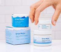 Box of Ursa Major Forest Alchemy Eye Cream on top of a jar of Alpine Rich Cream next to the jars of these products stacked on top of each other as a hand is grabbing the jar of Forest Alchemy Eye Cream in a white bathroom.