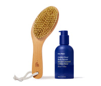 Wooden dry brush with a rope handle next to blue bottle of Ursa Major Golden Hour Body Serum on a white background