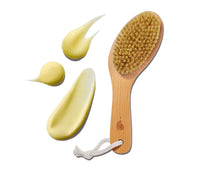 Wooden dry brush with bristles and Ursa Major bear logo next to droplets of yellow Body Serum on a white background
