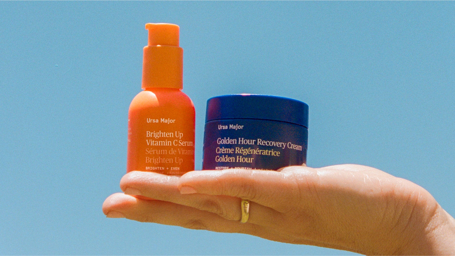 Hand holding Ursa Major Brighten Up Vitamin C Serum and Golden Hour Recovery Cream against a clear blue sky.
