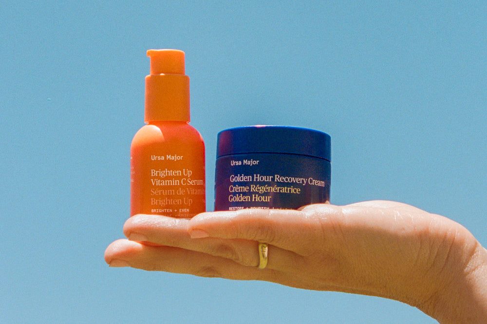 Hand holding Ursa Major Brighten Up Vitamin C Serum and Golden Hour Recovery Cream against a clear blue sky.