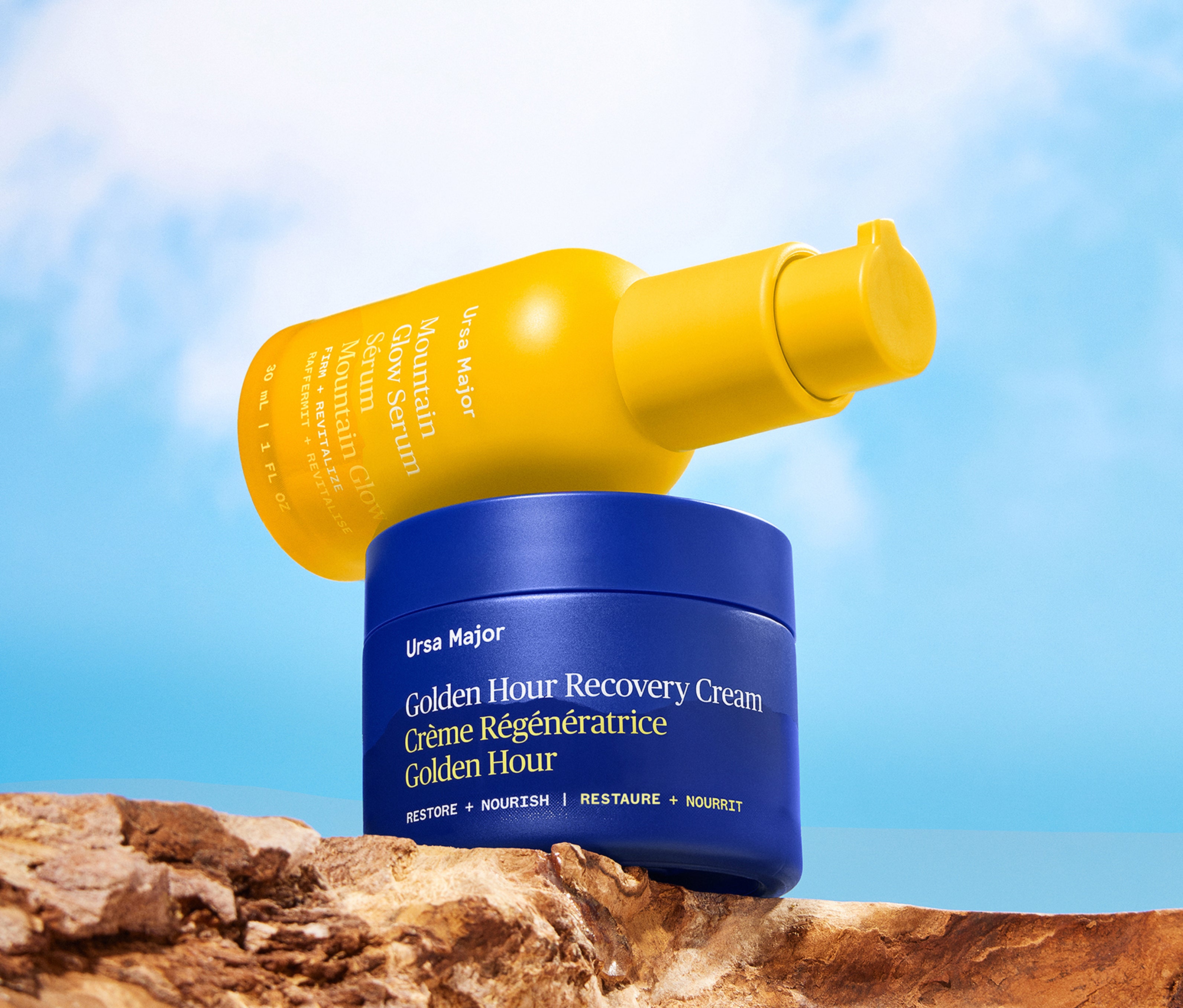 Yellow bottle of Mountain Glow Serum resting atop blue jar of Golden Hour Recovery Cream on a wooden surface with a blue sky background