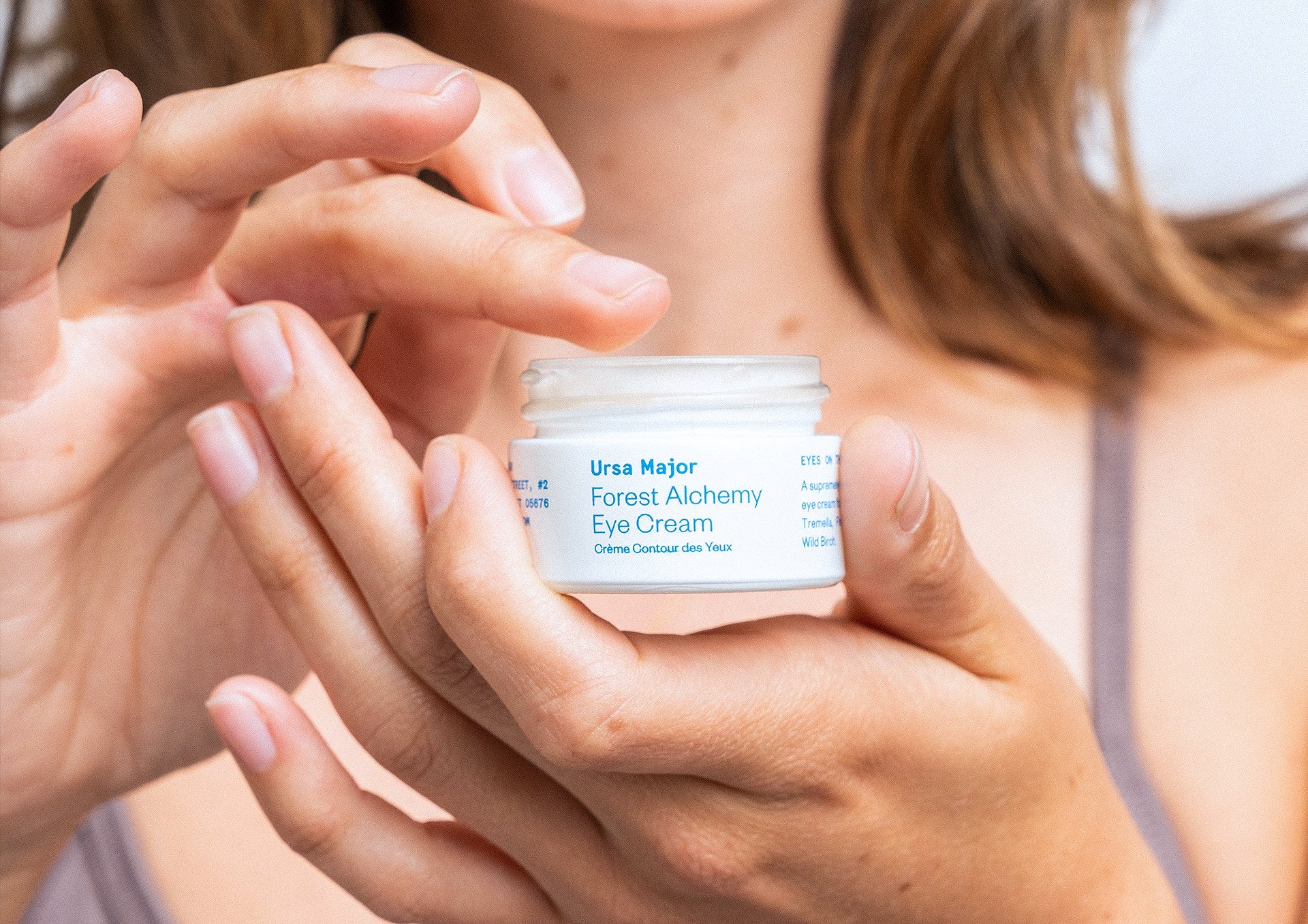 Close-up of a person holding an open jar of Ursa Major Forest Alchemy Eye Cream sticking their finger in it.