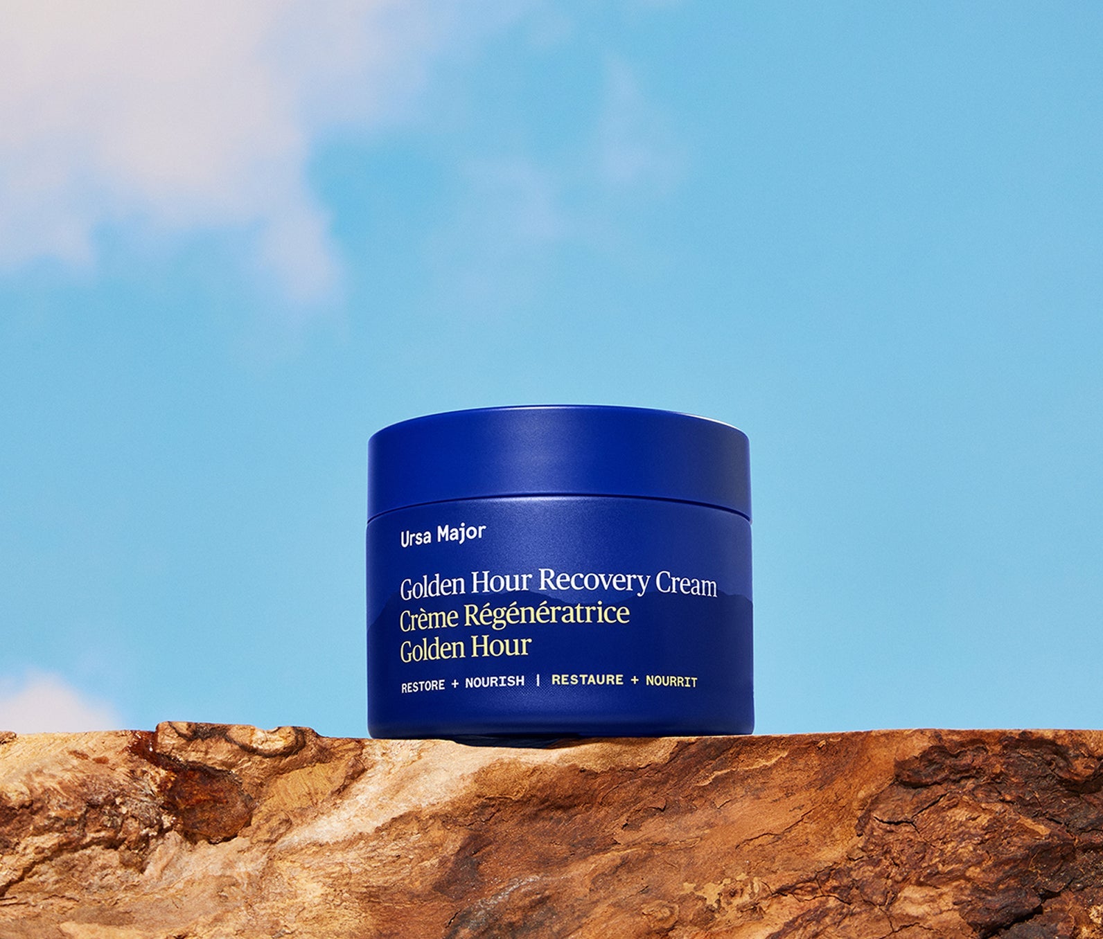 Blue jar of Ursa Major Golden Hour Recovery Cream on a wooden surface with a blue sky background