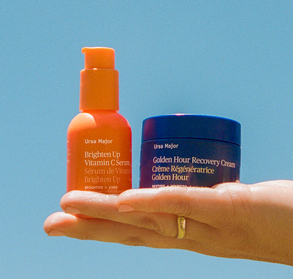 Hand holding Ursa Major Brighten Up Vitamin C Serum and Golden Hour Recovery Cream against a clear blue sky.