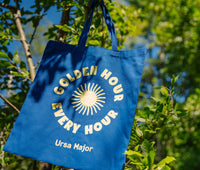 Blue tote bag with 'Golden Hour Every Hour Ursa Major' text and sun graphic hanging among trees.