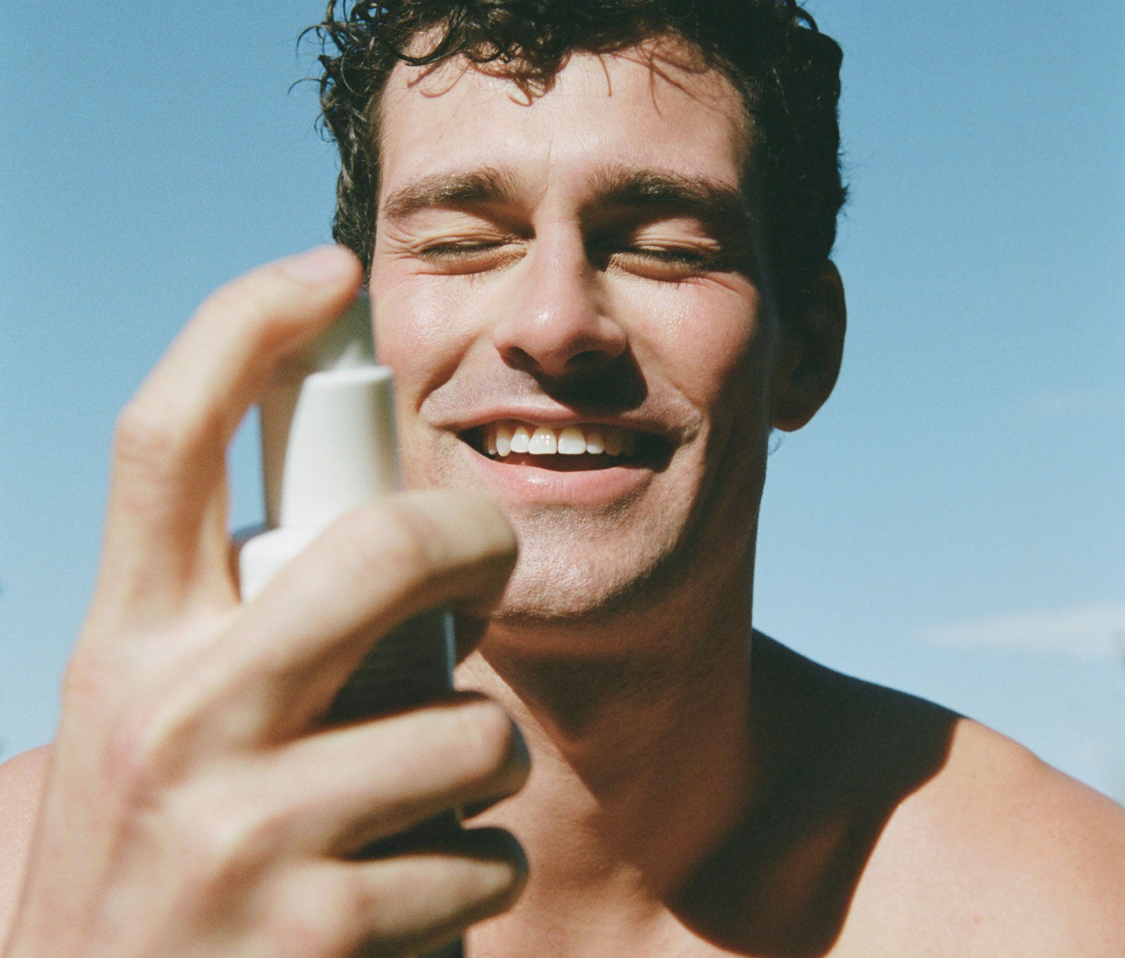 Smiling man refreshing his skin with a spray of tonic for his face, enjoying the cooling effects of the Ursa Major Essential Face Tonic.