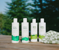 Ursa Major Perfect Zen Body Lotion, Citrus Riot Body Wash, Go Easy Conditioner, and Go Easy Shampoo sitting on top of a picnic table outside with a green and white checkered towel next to them.