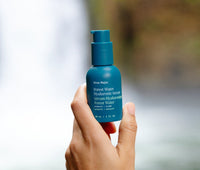 Hand holding a jar of Forest Water Hyaluronic Serum outside in front of a waterfall.