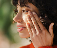 Profile of a woman looking to the left with sun on her face. Her hand in on her cheek rubbing in skincare.