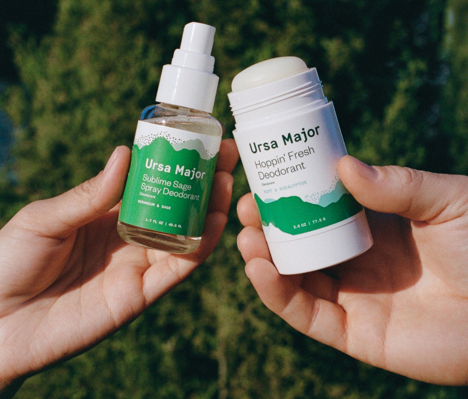 One hand holding Ursa Major Sublime Sage Spray Deodorant  next to a different hand holding Ursa Major Hoppin' Fresh Deodorant outside.