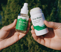 One hand holding Ursa Major Sublime Sage Spray Deodorant  next to a different hand holding Ursa Major Hoppin' Fresh Deodorant outside.