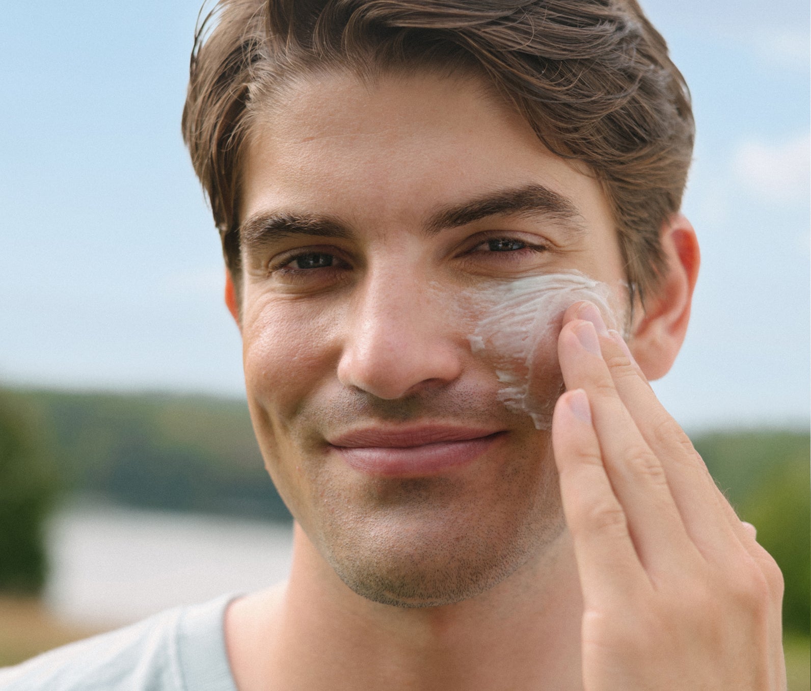 Smiling man applying cream to his cheek outdoors with a blurred natural background