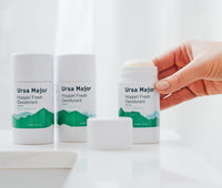 Three tubes of Ursa Major Hoppin' Fresh Deodorant on a white countertop  The right tube is open and the cap is to the side.  A hand is picking up the open tube.