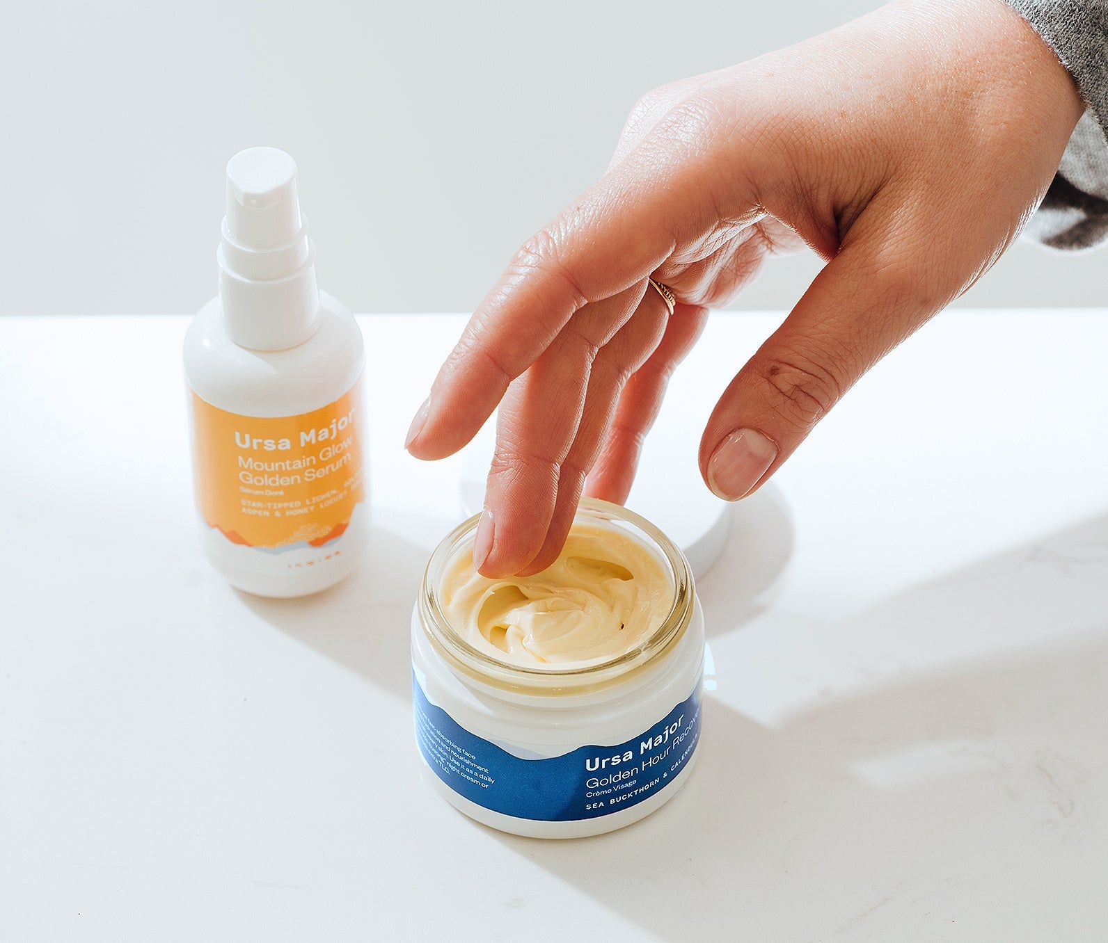 Ursa Major Mountain Glow Golden Serum bottle next to an open jar of Golden Hour Recovery Cream.  Hand is going to grab some of the Golden Hour Recovery Cream.