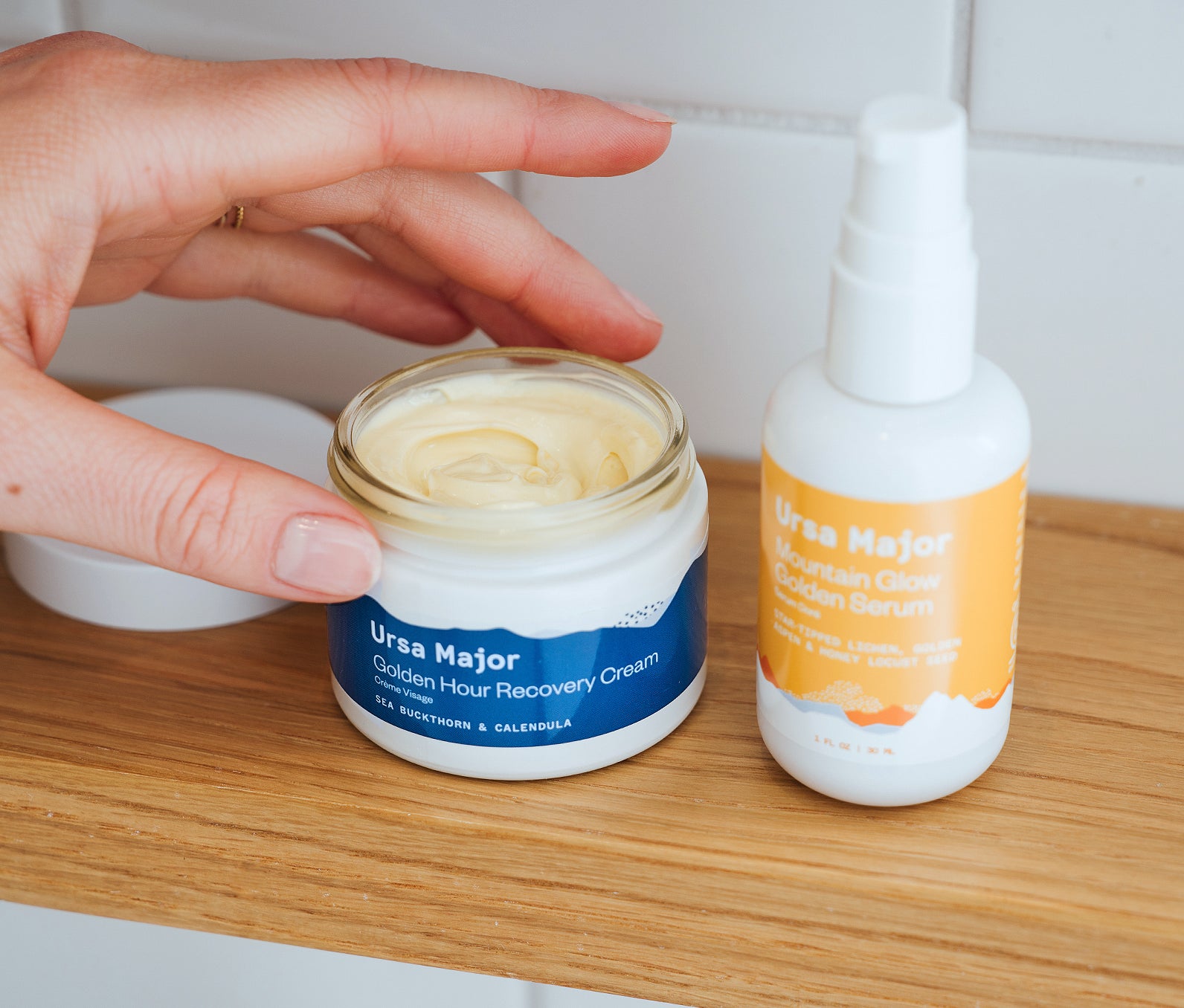 Open jar of Ursa Major Golden Hour Recovery Cream next to a bottle of Mountain Glow Golden Serum on a wooden counter with a white tiled wall behind them. Hand is going to grab some of the Golden Hour Recovery Cream.