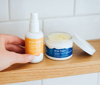 Open jar of Ursa Major Golden Hour Recovery Cream next to a bottle of Mountain Glow Golden Serum on a wooden counter with a white tiled wall behind them. Hand is going to grab the jar of Mountain Glow Golden Serum.