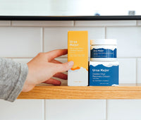 Hand placing a box of Ursa Major Mountain Glow Golden Serum next to a jar of Golden Hour Recovery Cream on top of the box of Golden Hour Recovery Cream on a wooden shelf.
